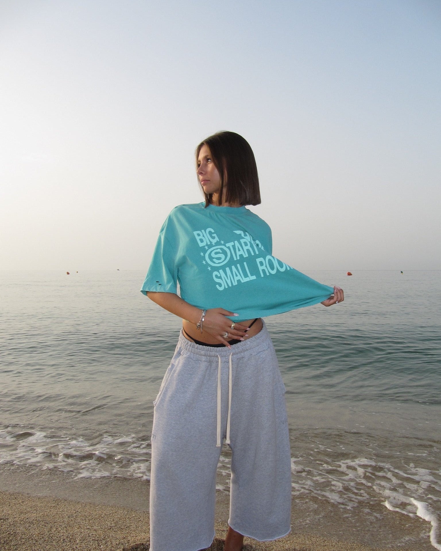 Model on the beach wearing the green Dreamer Green T-Shirt and grey Yellow Star Gray Pant, from Summer in Colors — Drop 02.